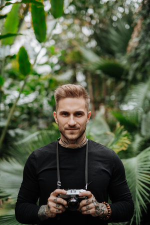 Handsome man with tattoos standing in botanical garden, in the middle of lush green foliage, holding camera and taking photography.の写真素材