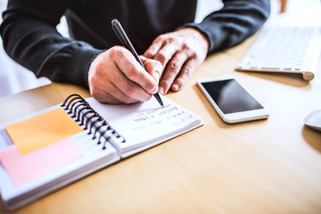 Close up of man writing notes in planner, working at desk from home. Telecommuting and home office.の写真素材