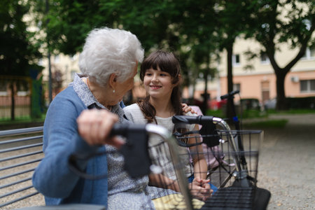 Grandma with walker spending time with granddaughter in city park, sitting on bench at children...