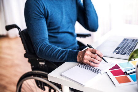 Close up of man in wheelchair working from home office, writing notes in planner. Telecommuting and home office.の写真素材