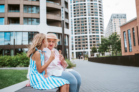 Grandma and granddaugter eating ice cream in the city, sitting on bench. Girl spending time with...