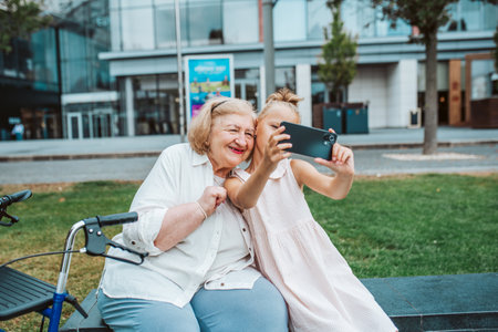 Grandma taking selfie with granddaugter sitting on bench. Girl spending time with senior grandmother, during summer break, after school.の写真素材