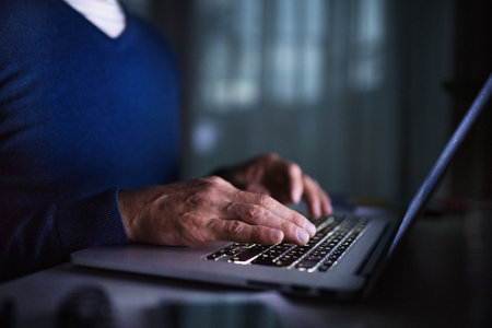 Close up on man working from home office at evening on laptop. Screen glow illuminates mans face. Telecommuting and home office.の写真素材