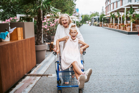Girl sitting on rollator, grandma pushing her, laughing. Granddaughter spending time with senior grandmother, during summer break, after school.の写真素材
