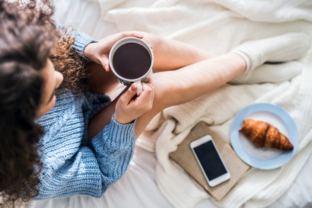 Morning cup of coffee. Beautiful young woman with curly hair sitting on bed in her room, drinking hot coffee.の写真素材