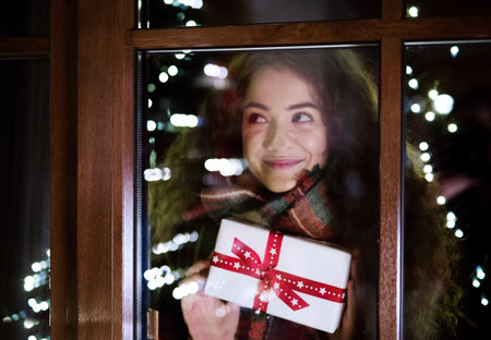 Student is coming home from university for the Christmas holidays, carrying bunch of Christmas presents and looking inside through window.の写真素材
