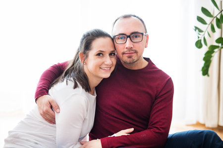 Husband and wife are sitting on the floor in living room, hugging and looking at the camera.
