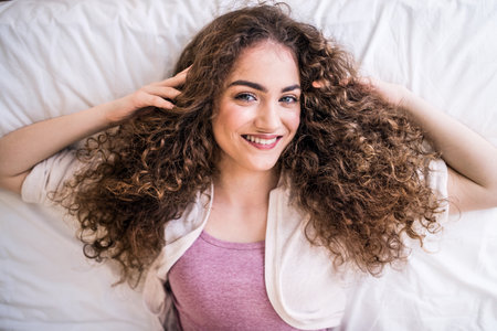 Portrait of beautiful young woman with a curly hair lying on her back on bed. Top view shot.
