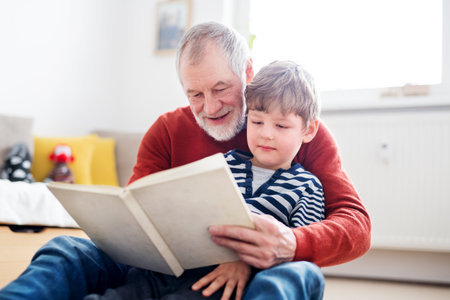 Grandfather spending time with grandson, reading storybook together, sitting on carpet.の写真素材