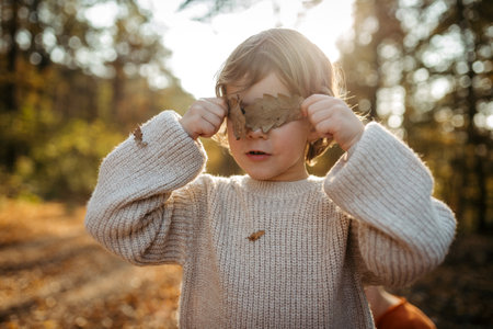 Portrait of cute girl standing in the middle of autumn forest, covering her eyes with dry leaves.の写真素材