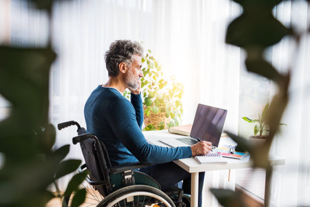 Handsome man in wheelchair working from home office, making phone call and writing notes in planner....
