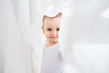 Cute little boy is standing on indoor windowsill, hiding behind the curtains.の写真素材