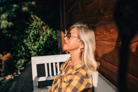 Beautiful older woman is sitting in front of her cabin, enjoying autumn sun rays on her face.の写真素材