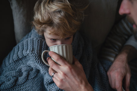 Father helping young son to drink warm tea from cup. Sick boy with cold lying under blanket and drinking herbal tea.の写真素材