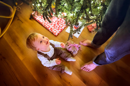 Little baby boy sitting by Christmas tree, looking up at his father. Cute boy is surprised and...
