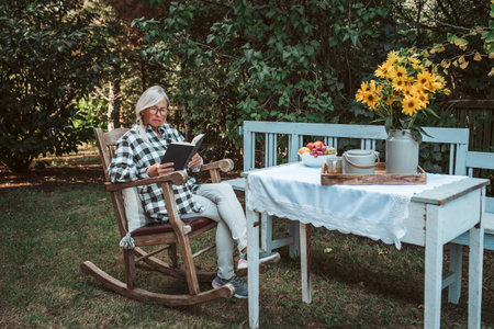 Beautiful older women is sitting in rocking chair in the garden and reading book. Female senior is enjoying a peaceful moment at home, savoring her freedom.の写真素材