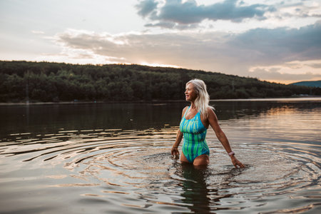 Cold water swimming for elderly women. Senior sporty women standing in lake during cold evening, looking at setting sun.の写真素材