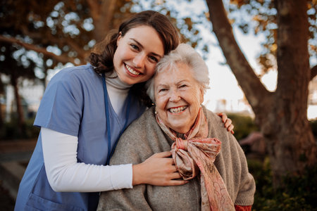 Portrait of nurse and elderly woman on walk in the park during warm autumn day. Young caregiver spending time with senior patient.の写真素材