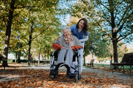 Disabled elderly woman in wheelchair on walk in the park during warm autumn day. Young nurse pushing wheelchair, talking with senior woman.の写真素材
