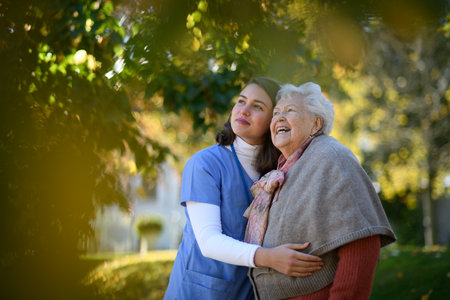 Portrait of nurse and elderly woman on walk in the park during warm autumn day. Young caregiver spending time with senior patient.の写真素材
