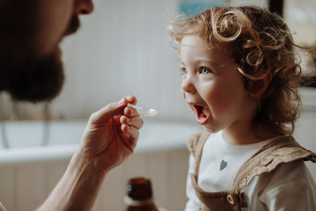 Sick girl with fever and cold at home. Father giving his daughter medicine on spoon, cough syrup or fever medicine.の写真素材