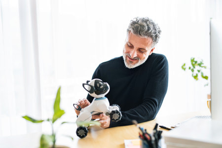 Handsome man working from home office, sitting at desk with computer and holding robotic dog. Telecommuting and home office.の写真素材