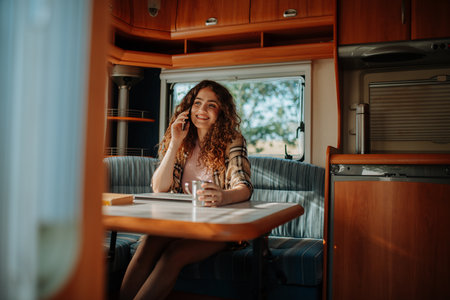 Young beautiful woman is on a camping trip in nature, sitting in her caravan making phone call. Caravan as homeoffice, working remotely.の写真素材