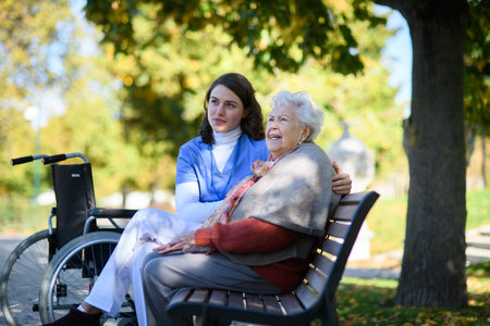 Portrait of nurse and elderly woman sitting on bench in park during warm autumn day. Young caregiver spending time with senior patient.の写真素材