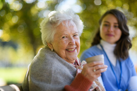 Elderly woman enjoying cup of coffee with her nurse in the park during warm autumn day. Young...