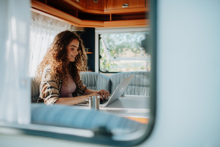 Young beautiful woman is camping in nature, sitting in her caravan and working on a notebook. Caravan as homeoffice, working remotely. Through the window.の写真素材