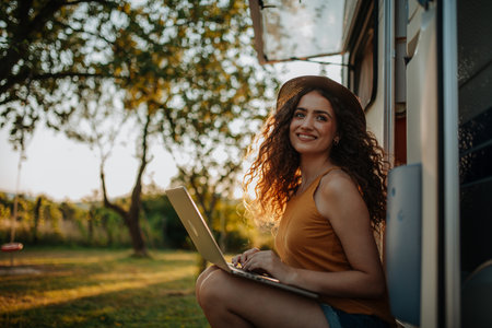 Young beautiful woman is on a camping trip in nature, sitting in front of caravan with notebook on knees. Caravan as homeoffice, working remotely.の写真素材