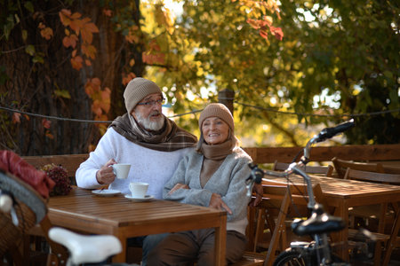 Elderly married couple sitting on a coffee shop terrace on beautiful autumn day, enjoying cup of coffee in the autumn atmosphere.の写真素材