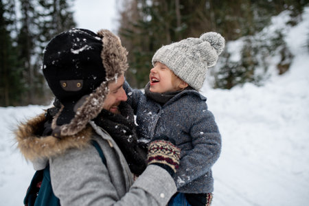 Father standing in the middle of snowy forest holding young son in arms. Dad and boy enjoying winter holiday in the mountains.の写真素材