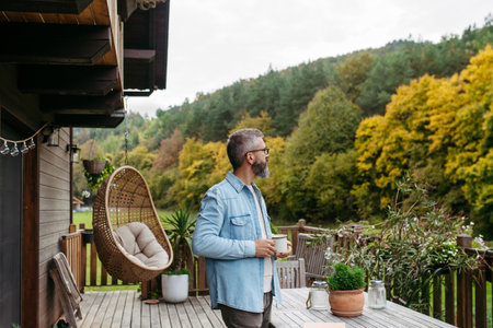 Man is enjoying cup of warm tea, coffee, having relaxing moment at home, standing on patio and looking at autumn nature around him.の写真素材