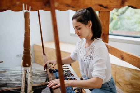 Young businesswoman sitting at a wooden loom, weaving a rug from old fabrics. Upcycling in business and circular economy.の写真素材