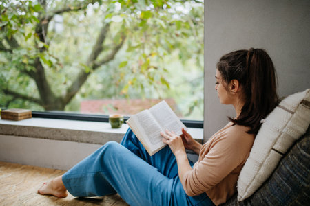Hygge moment for beautiful woman sitting by a large window with a book. Woman is enjoying time for herself, reading.の写真素材