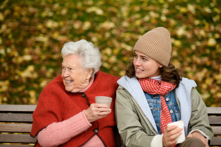 Portrait of a granddaughter and grandmother sitting on bench in autumn park, drinking coffee and laughing together.の写真素材
