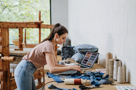 Young businesswoman working on laptop on workbench, standing by wooden loom. Upcycled textile, small...