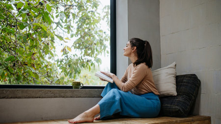 Hygge moment for beautiful woman sitting by a large window with a book. Woman is enjoying time for herself, reading.の写真素材
