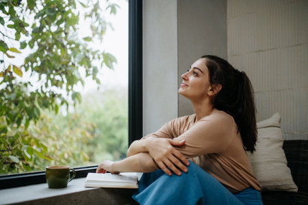 Hygge moment for beautiful woman sitting by a large window with a book. Woman is enjoying time for herself, reading.の写真素材