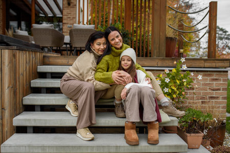 Mature daughter, grandmother and granddaugter sitting on wooden stairs, hugging. Mothers day and...