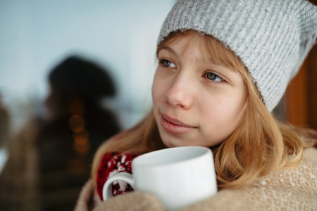 Pensive teen girl bundled in thick blanket holding cup of hot tea standing on balcony during winter day.の写真素材