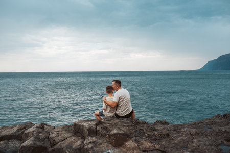Rear view of father and son fishing in the sea. Sitting on rock holding fish rod.
