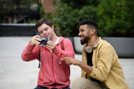 Young man with Down syndrome and mentoring friend spending time together, using camera outdoor.の写真素材