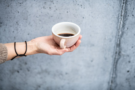 Close up of hand holding cup with coffee in front of concrete wall.の写真素材
