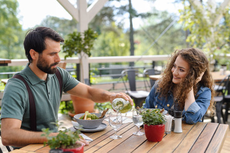 Young couple at date in restaurant, sitting on restaurant terrace. Boyfriend pouring water in...