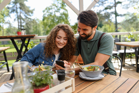 Friends sitting on restaurant terrace, meeting after long time, looking at old photos in smartphone. Friends enjoying springtime, having lunch outdoors.の写真素材