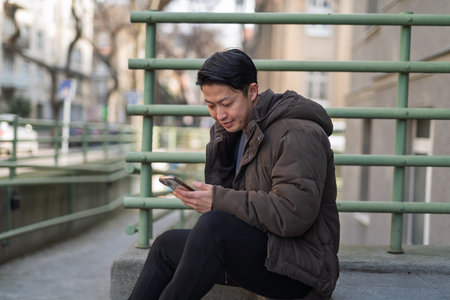 Young Asian man sitting on street ledge scrolling on smartphone