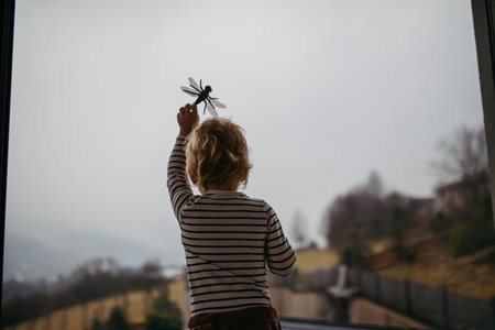 Cute toddler boy is playing with dragonfly toy, standing by window.の写真素材