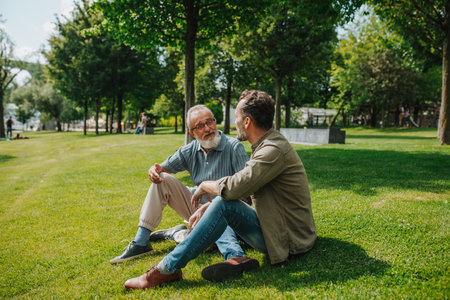 Father and adult son sitting on grass in park, sharing a story from the past.の写真素材